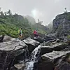 To turgåere går over store steiner ved et lite fossefall i et bratt, grønt fjellandskap.
