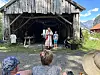 Folk sitter og står foran en gammel løe, med to personer i midten og fjell i bakgrunnen.