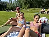 To jenter sitter og ligger på et teppe på gresset ved en strand mens folk slapper av i bakgrunnen.