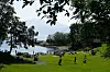 Folk sitter og går på en gressbakke ved vannet, med trær som rammer inn utsikten mot fjorden.