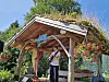 En mann spiller gitar og synger på en liten scene med torvtak og hengende blomster.