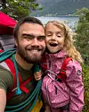 En mann og et barn smiler tett sammen utendørs med fjord, fjell og vann i bakgrunnen.