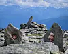 To personer smiler fra åpninger mellom steiner på en fjelltopp med fjellandskap i bakgrunnen.