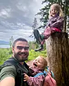 En mann tar selfie med to små barn utendørs ved en stor trestubbe i skogen.