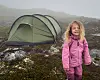 Et barn i rosa jakke står ved siden av et grønt telt på lyngkledd fjellterreng i tåke.