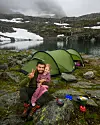 En mann og et barn sitter foran et grønt telt ved et fjellvann med snøflekker i fjellsiden bak.