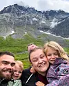 En smilende familie står tett sammen foran et bratt fjell med grønne og steinete sider.