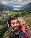 En mann og et barn tar en selfi på en fjelltur med stor tursekk og fjordutsikt bak.