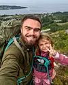 En mann og et barn tar en selfie utendørs med fjord og øyer i bakgrunnen.