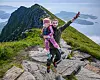 En mann bærer et barn på en steinete fjelltopp med fjord og øyer i bakgrunnen.