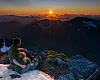 To personer sitter på en snøkant med utsikt mot solnedgang over fjellene.