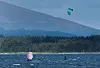 To kitesurfere på vannet med turkise og rosa drager foran en skogkledd kyst og et fjell.