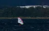 En kiteboarder med fargerik seilglider på mørkt vann foran en skogkledd kyst.
