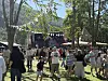 Mennesker står og går på en gresslette foran en liten scene i et parkområde med trær.