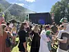 Folk står samlet på gresset foran en stor utendørsscene i solskinn, med fjell og trær i bakgrunnen.
