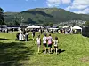 Personer står og går på en grønn gresslette med telt, boder og fjell under en blå himmel.