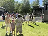 Mange mennesker står på gresset foran en scene i en park på en solrik dag.