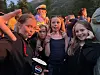 Tre barn står tett sammen ute, med fjell og flere personer i bakgrunnen ved solnedgang.