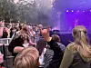 Folk sitter og står foran en scene med lilla lys under en utendørskonsert.