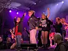 Flere personer står og danser på en scene med lilla lys og publikum foran.