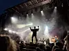 En person står med armene hevet på en opplyst scene med bandutstyr og kraftige lysstråler.