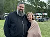 To personer står sammen på en gresslette med arrangementstelt og mennesker i bakgrunnen.