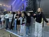 En gruppe ungdommer står foran en stor scene med lys og musikere i bakgrunnen.