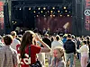 Folk står tett foran en utendørsscene der musikere opptrer under hengende scenelys.