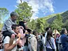 En gruppe mennesker står samlet utendørs foran trær og fjell under blå himmel.