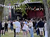Folk står på gresset foran en utendørs scene med lys, flagg og et KOFF-banner.