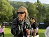 En kvinne med solbriller og svart jakke står ute i et grønt festivalområde med andre mennesker i bakgrunnen.