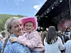 Ei kvinne holder et barn med rosa hatt foran en utendørsscene med publikum og røyk i bakgrunnen.