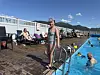 Utendørs basseng med badegjester, solsenger og en person ved stigen i forgrunnen.