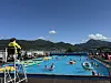 Utendørsbasseng med mange badende, livredderstol og fjell i bakgrunnen under blå himmel.