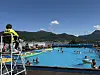 Utendørs svømmebasseng med mange badende, badevakt i stol og fjell i bakgrunnen.