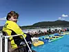 Person sitter på stol ved et utendørsbasseng med badeleker og folk i vannet, med fjell og blå himmel i bakgrunnen.