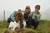 Tre barn sitter tett sammen med flere sauer på en grønn, gresskledd bakke.