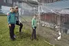 Tre personer står på gress ved et gjerde, med en hvit gås og et lite uthus i bakgrunnen.