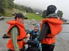 Tre personer i oransje redningsvester står ved vannet med grønne åser og små bygninger i bakgrunnen.