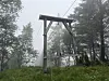 Gamle taubanestolper står i en bratt, grønn skogkledd fjellside i tett tåke.