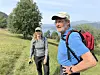 To personer står på en grasdekt sti med fjell og landskap i bakgrunnen.