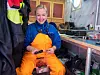 En smilende person i orange verneklær sitter i en fabrikk og holder en snøkrabbe.