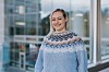 En kvinne smiler foran store vinduer med utsikt mot et moderne bygg og vann i bakgrunnen.