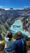 Et par sitter på en fjellkant og ser ut over en turkis innsjø mellom bratte fjell.