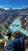 Et par sitter på en fjellkant og ser ut over en turkis innsjø mellom bratte fjell.