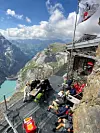 Flere personer sitter og hviler på en fjellterrasse ved en steinhytte med utsikt over fjell og innsjø.