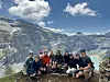 En gruppe mennesker sitter og står på en steinrygg foran snødekte fjell.