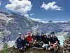En gruppe mennesker sitter og står samlet foran høye fjell, med snøfelt, innsjø og blå himmel i bakgrunnen.