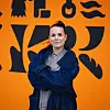 Kvinne står foran en oransje vegg med svarte abstrakte symboler, med armene i kors.