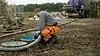 En person i oransje arbeidsbukse arbeider ved en kum med slange på en gjørmete gårdsplass.